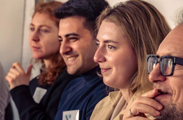 Vier Personen sitzen in einer Reihe nebeneinander und schauen zum linken Bildrand. 