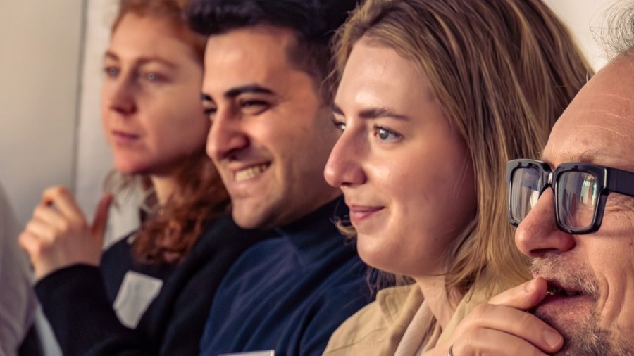 Vier Personen sitzen in einer Reihe nebeneinander und schauen zum linken Bildrand. 