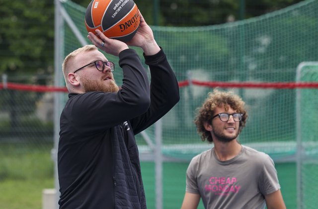 Ein Mann hält einen Basketball in beiden Händen, um ihn in die Luft zu werfen. Ein anderer Mann ist im Hintergrund zu sehen und schaut in die Flugrichtung des Balls. 