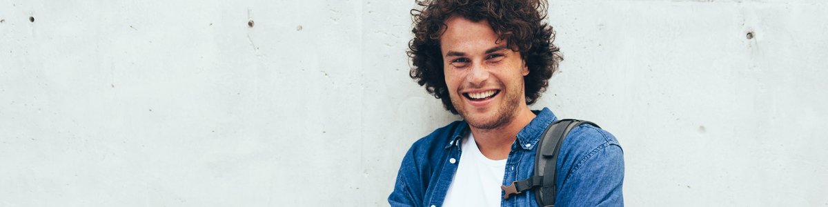 Lächelnder junger Mensch mit Rucksack auf dem Rücken, der in Freizeitkleidung vor einem Betongebäude auf einer Stadtstraße steht