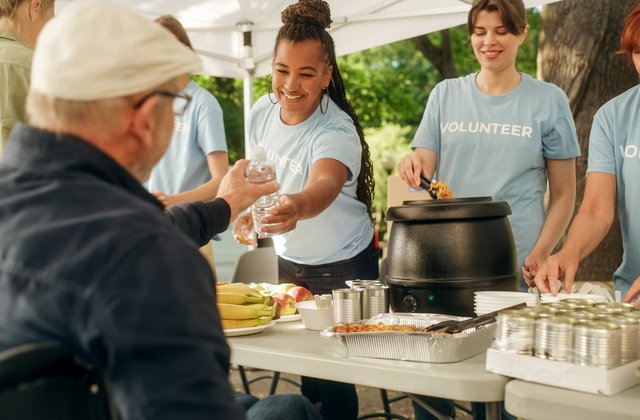 Ein Team von Freiwilligen hilft in einer Lebensmittelbank und verteilt an einem sonnigen Tag in einem Park Lebensmittel. Ein Mensch mit Behinderung, der einen Rollstuhl benutzt, nimmt die Mahlzeit entgegen