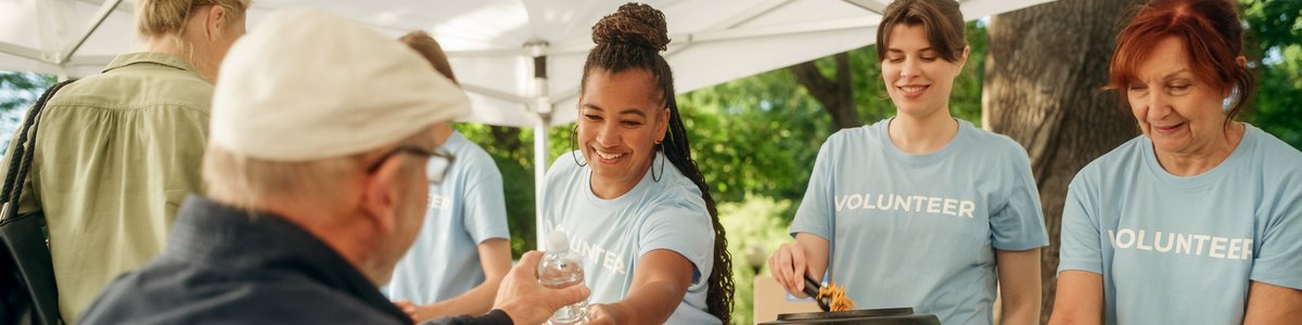 Ein Team von Freiwilligen hilft in einer Lebensmittelbank und verteilt an einem sonnigen Tag in einem Park Lebensmittel. Ein Mensch mit Behinderung, der einen Rollstuhl benutzt, nimmt die Mahlzeit entgegen