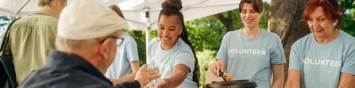 Ein Team von Freiwilligen hilft in einer Lebensmittelbank und verteilt an einem sonnigen Tag in einem Park Lebensmittel. Ein Mensch mit Behinderung, der einen Rollstuhl benutzt, nimmt die Mahlzeit entgegen