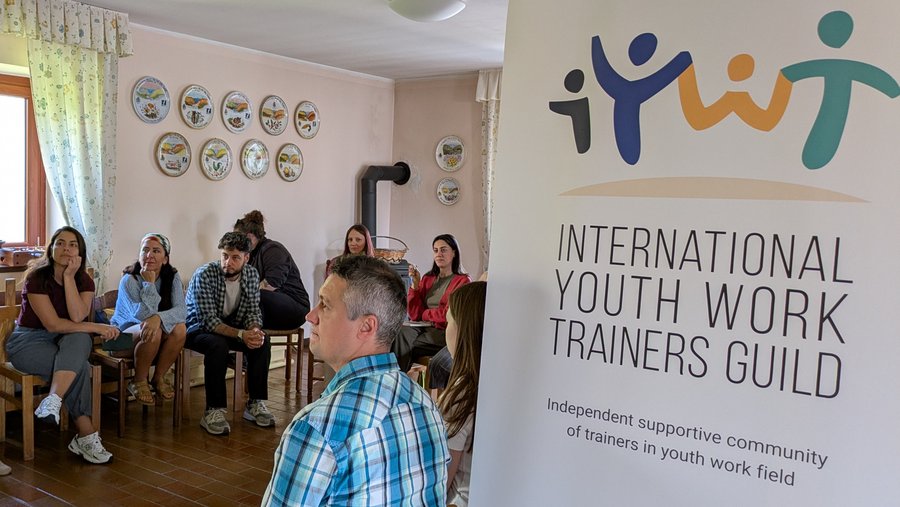 The picture shows a Pull-up banner with the text "International Youth Work Trainers Guild - Independent supportive community of trainers in youth work field". Next to the banner are people sitting together in a group.