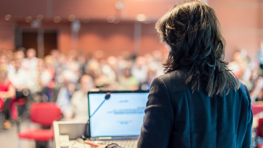 Eine Person hält einen Vortrag auf einer Konferenz. Unbekannte Personen im Publikum des Konferenzsaals