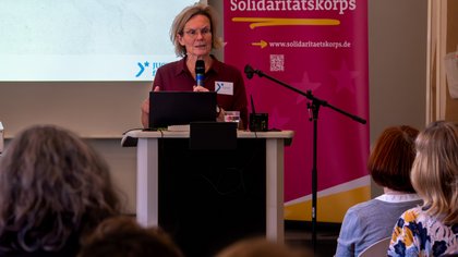 Eine Person steht hinter einem Rednerpult spricht in ein Mikrofon. Im Hintergrund sieht man den Ausschnitt einer Leinwand und einen Roll Up Banner. 