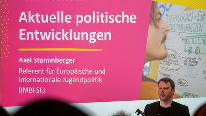 Powerpoint Präsentation auf einem Beamer. Davor steht ein Sprecher. Köpfe des Publikums im unteren Bildrand. 