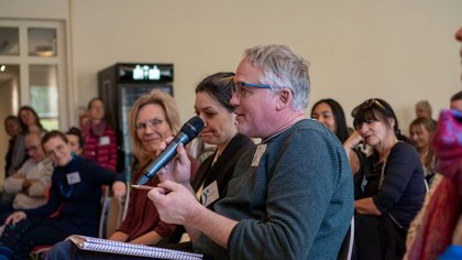 Mehrere Personen sitzen auf Stühlen in einem Raum, einer hält ein Mikrofon und spricht. 
