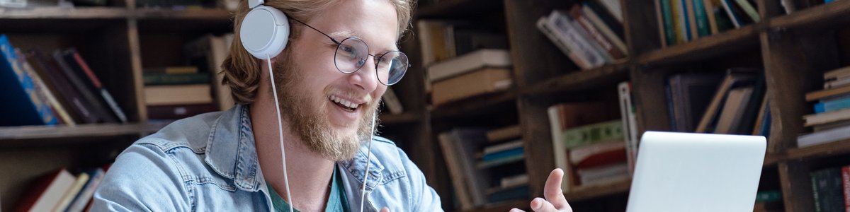 Fröhlicher junger Mnesch trägt Kopfhörer, führt ein Video-Gespräch in der Bibliothek und schaut dabei auf den Laptop-Bildschirm