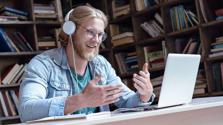 Fröhlicher junger Mnesch trägt Kopfhörer, führt ein Video-Gespräch in der Bibliothek und schaut dabei auf den Laptop-Bildschirm