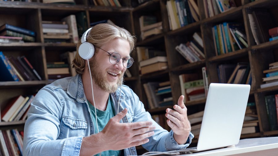 Fröhlicher junger Mensch trägt Kopfhörer, führt ein Video-Gespräch in der Bibliothek und schaut dabei auf den Laptop-Bildschirm