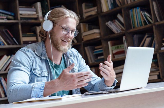 Fröhlicher junger Mnesch trägt Kopfhörer, führt ein Video-Gespräch in der Bibliothek und schaut dabei auf den Laptop-Bildschirm