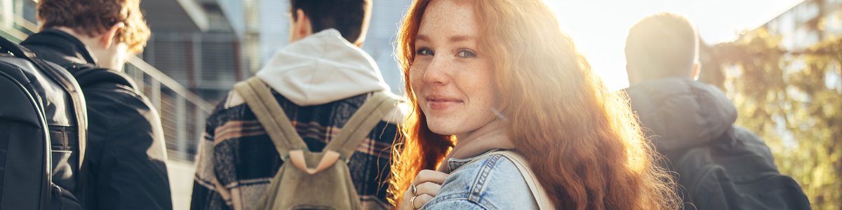 Drei junge Menschen gehen mit Rucksäcken und den Rücken zur Kamera eine Straße entlang. Eine junge Person mit Umhängetasche dreht sich lächelnd der Kamera zu
