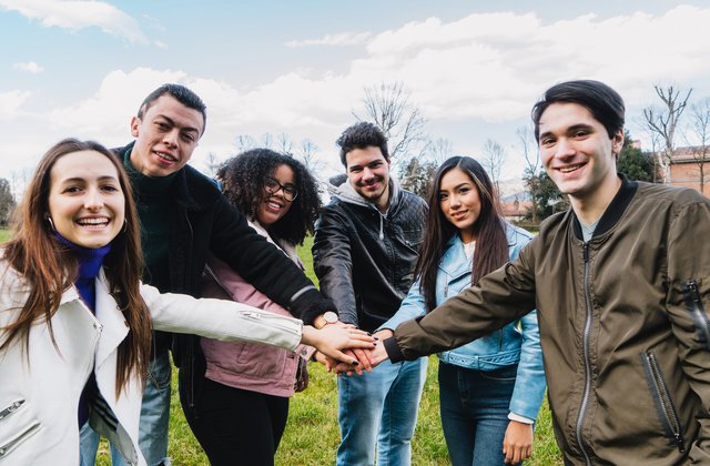 Eine Gruppe junger Menschen im Park fasst sich in der Mitte eines Kreises an den Händen und hat gemeinsam Spaß