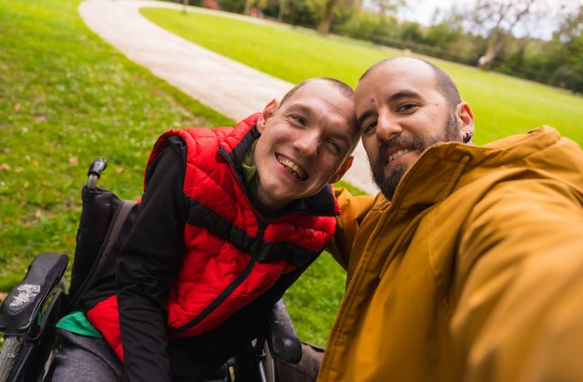 Einer Person mit Behinderung im Rollstuhl und eine andere Person machen ein Selfie auf einer Bank in einem öffentlichen Park in der Stadt; sie unterhalten sich und lachen