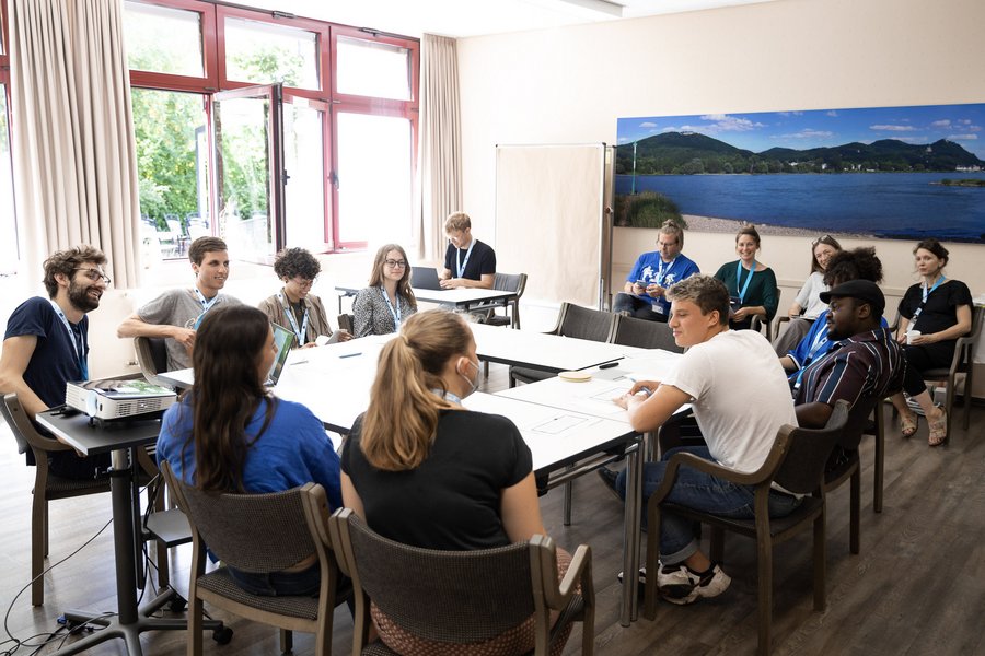 Das Bild zeigt Jugendliche, die in einem Raum an Tischen in U-Form sitzen und sich miteinander austauschen.