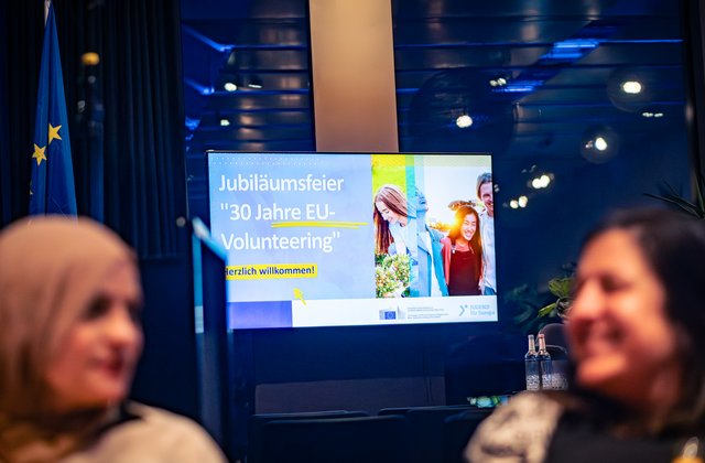 Ein Bildschirm zeigt den Text „Jubiläumsfeier 30 Jahre EU‑Volunteering – Herzlich willkommen“, seitlich ist eine EU‑Flagge zu sehen, im Vordergrund unscharfe Personen.