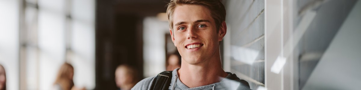 Junger Mensch steht auf dem Flur, an eine geflieste Wand gelehnt, und lächelt