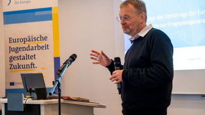 In der linken Bildhälfte steht ein Roll-up Banner hinter einem Tisch mit Laptop, in der rechten Bildhälfte steht ein Mensch hinter einem Mikrofon vor einer Leinwand. 