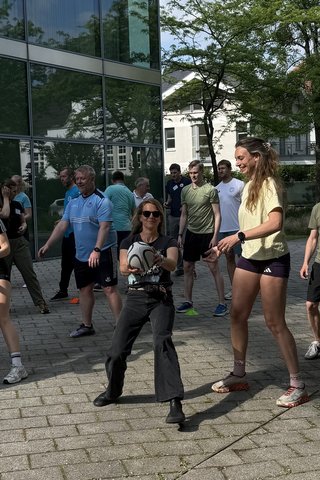 Menschen in Sportkleidungen stehen in zwei langen Reihen hintereinander und reichen sich einen Volleyball